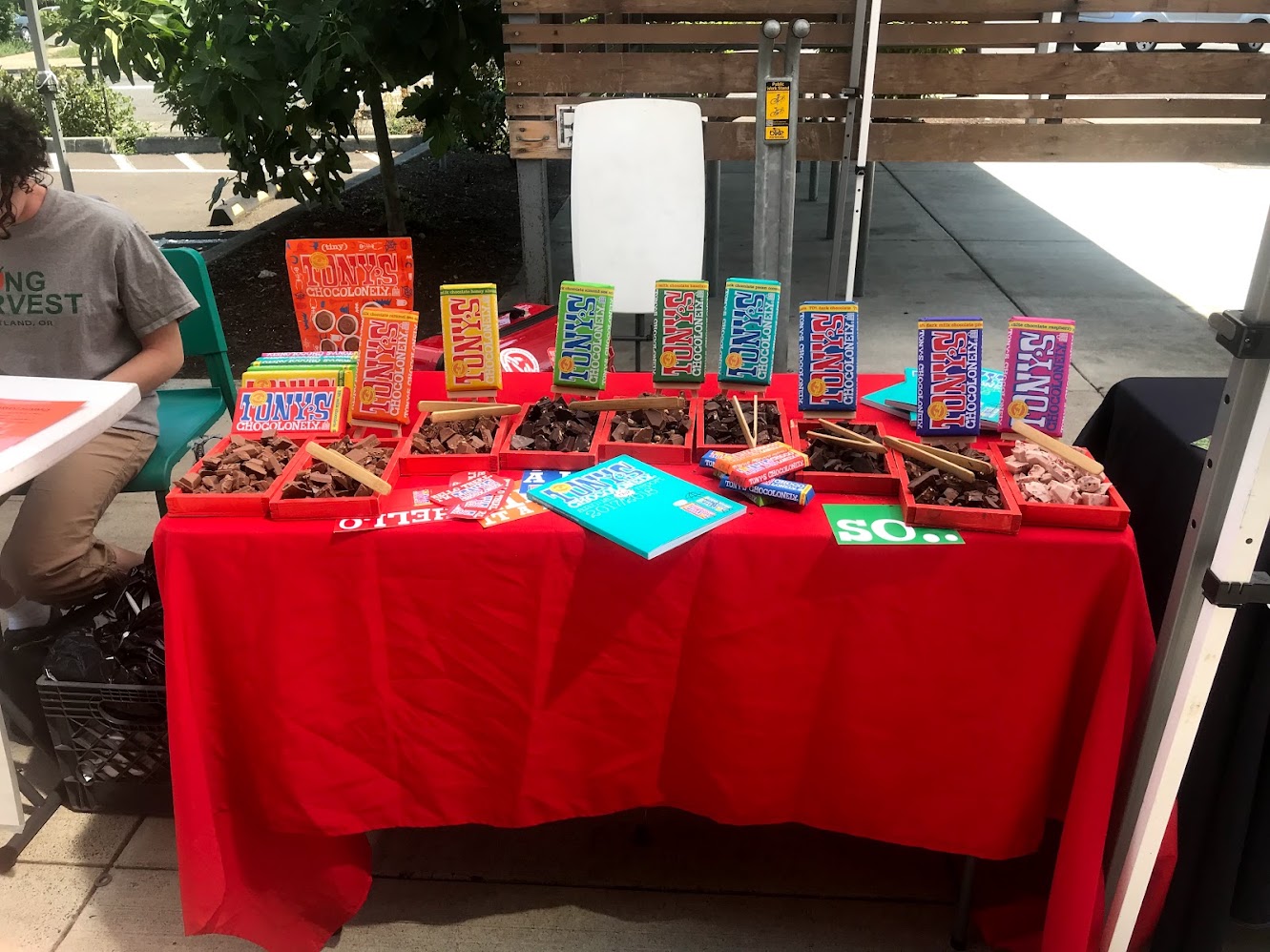 An outdoor pop-up table with a red cloth, Tony’s bars standing up, and bins of chocolate samples